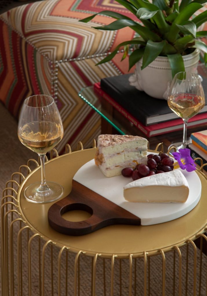 Dois pedaços de queijo e um cacho de uvas vermelhas sobre uma tábua arredondada. Ao lado, há uma taça com vinho branco. 