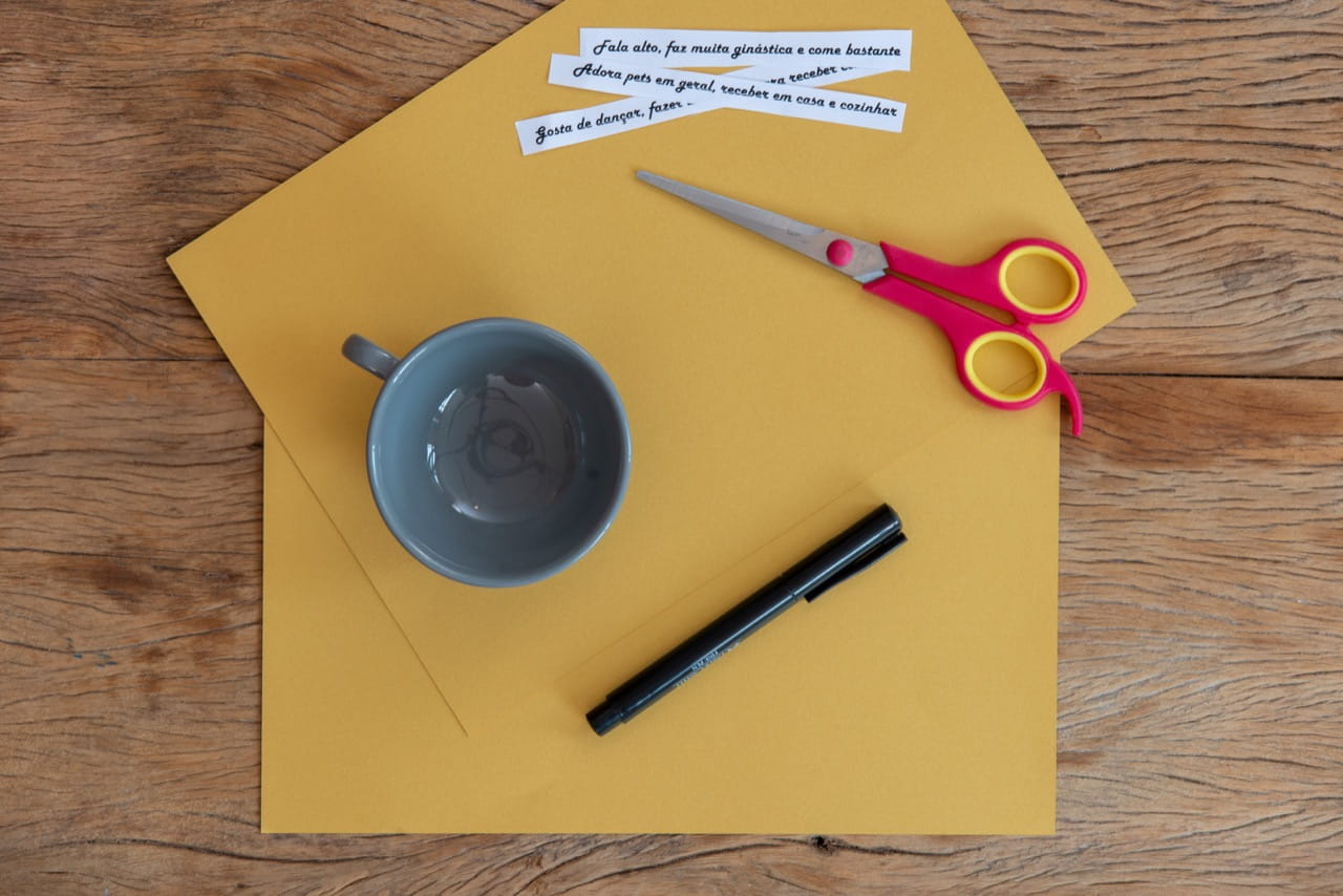 Material para o sorteio de amigo-secreto: duas folhas de papel amarelo mostarda, uma xícara cinza, caneta, tesoura e frases escritas em preto num papel branco.