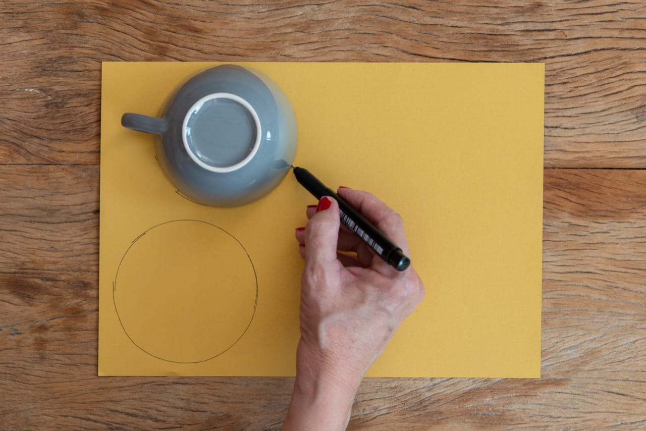 Xícara de boca para baixo sobre uma folha de papel. Uma mão feminina desenha o contorno com a caneta, criando círcúlos.