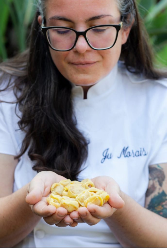 Retrato da chef Juliana Ferreira, mulher branca com longos cabelos castanhos e óculos de grau. Ela segura um punhado de massa fresca nas palmas das duas mãos em concha. 