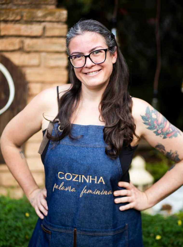 Retrato da chef Juliana Ferreira, mulher branca com longos cabelos castanhos, vestindo avental de cozinha jeans, com a frase "Cozinha, palavra feminina", bordada em amarelo.