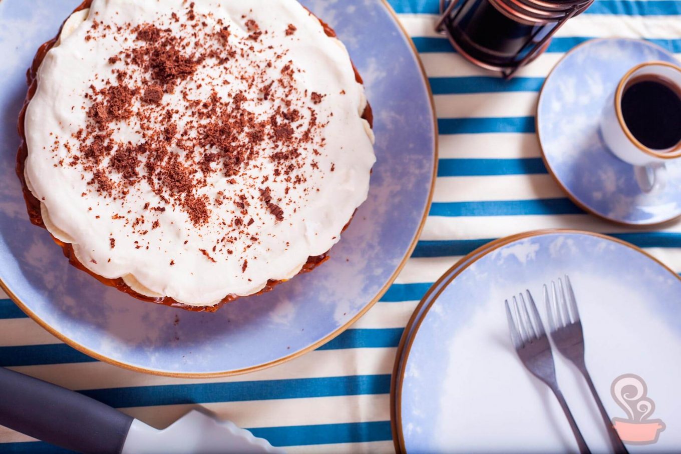 Foto feita de cima mostra uma torta com cobertura branca, prato de sobremesa e xícara de café