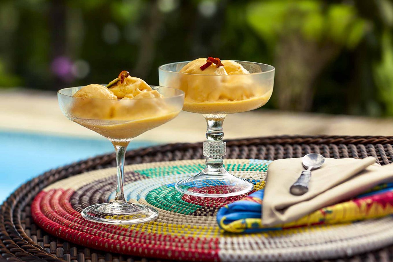 Duas taças de vidro contendo creme de manga. Elas estão sobre uma bandeja colorida, com um piscina ao fundo. 