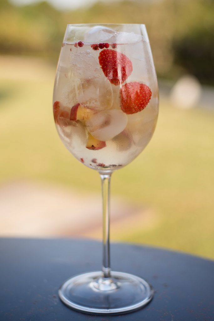 Taça de cristal com drink, gelo e frutas. Ao fundo aparece um jardim fora de foco.