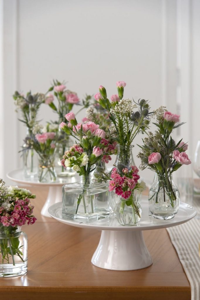 Várias garrafas com flores sobre um prato de bolo, branco, com pé. 