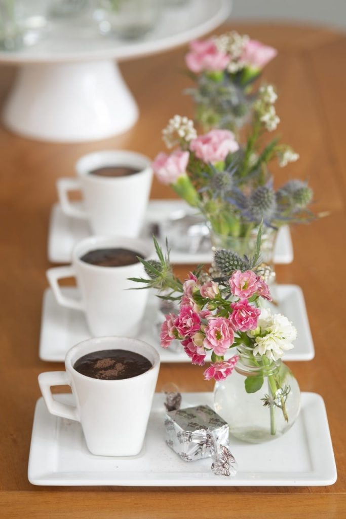 Três xícaras sobre pires retangulares, ambos de porcelana branca. Sobre cada pires há uma xícara, um bombom e um vasinho com flores. 