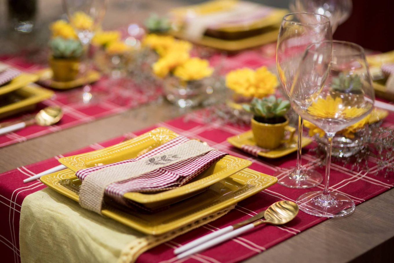 O xadrez vermelho e branco, presente nos caminhos de mesa e guardanapos, deixa a mesa com um ar campestre. Uma fita de juta, com um carimbo personalizado, abraça cada conjunto de pratos. Há, ainda um pano de prato amarelo embaixo deles, para criar mais um ponto de cor.