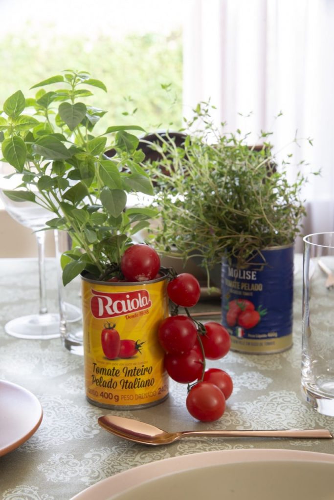 Duas latas de tomate pelado, uma com rótulo amarelo e outra com rótulo azul, contemdo ervas e tomates em rama. 