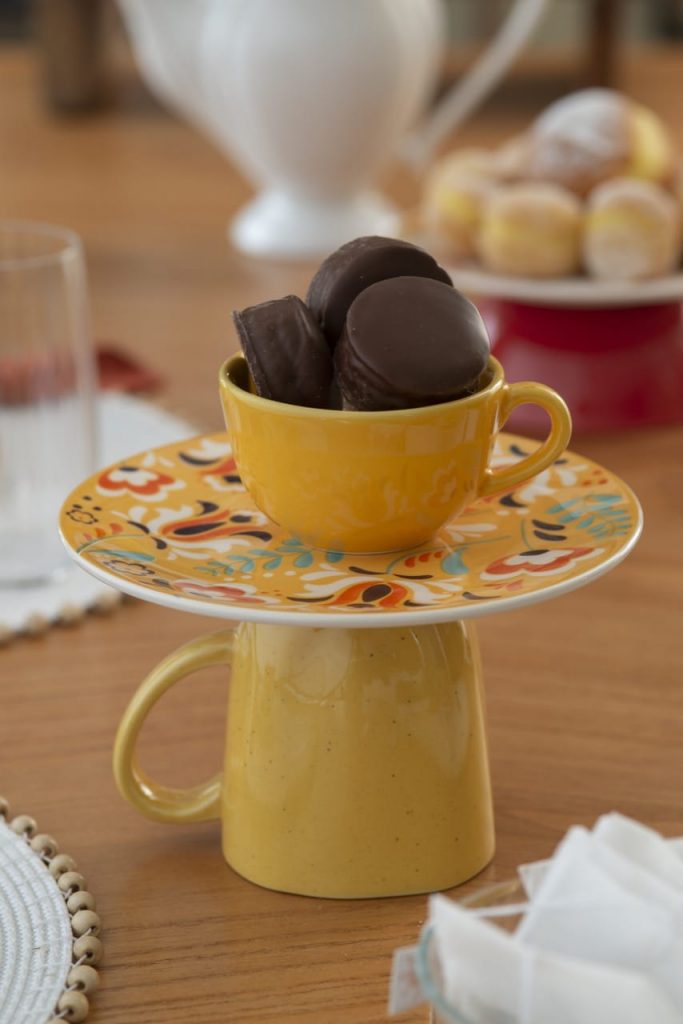 Xícara amarela contendo pães doces está sobre um prato de sobremesa apoiado numa caneca com a boca virada para baixo. 