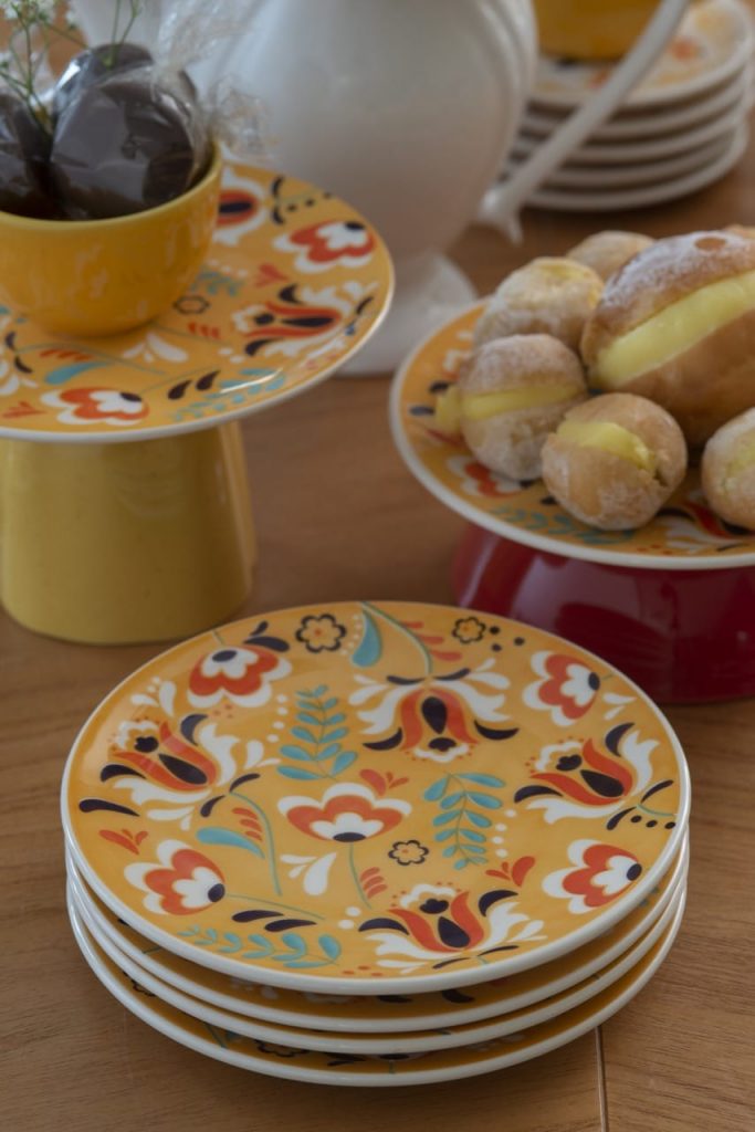 Na mesa polonesa, temos uma pilha de pratos amarelos com estampa de flores estilizadas em branco, laranja e marrom. 