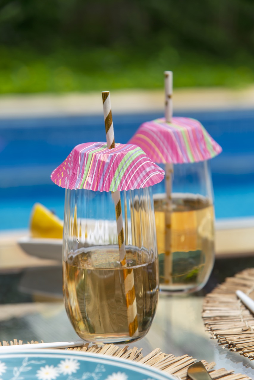 Dois copos com drink dourado, cobertos por forminha listradas em tons de rosa, verde e azul, com canudos de papel branco e dourado. Ao fundo, vemos uma piscina. 