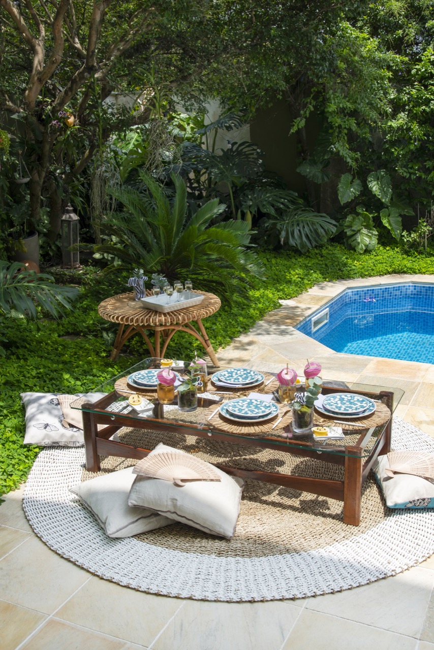 Mesa ao ar livre para almoço num jardim com muitas plantas. Pode-se ver parte de uma piscina., do lado direito.