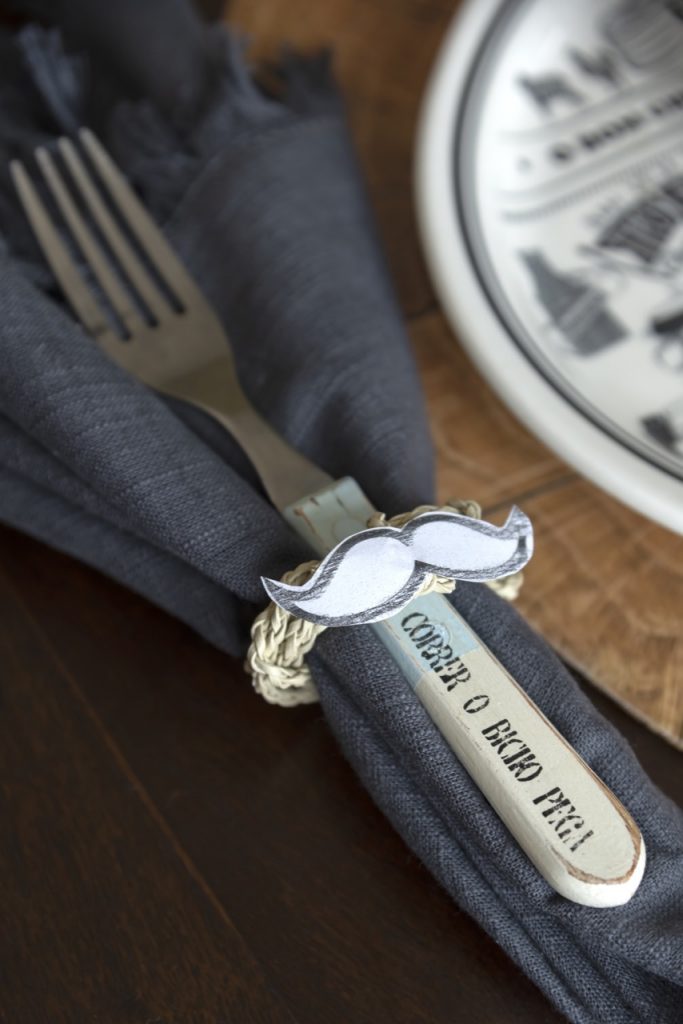 Na mesa posta, foto aproximada do anel de guardanapo com bigode. Dentro dele, temos um garfo e um guardanapo cinza. 