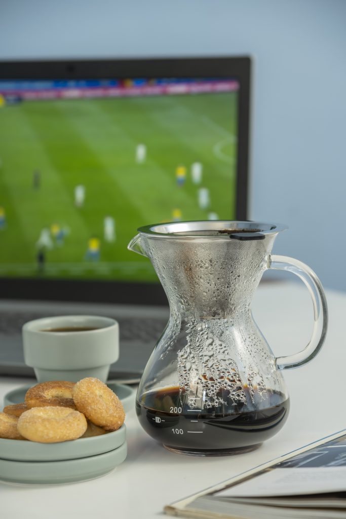 Passador de café de vidro e metal, contendo café. Do lado esquerdo, vemos dois pires empilhados com 4 biscoitos, e uma xícara de cafezinho sobre um pires. 