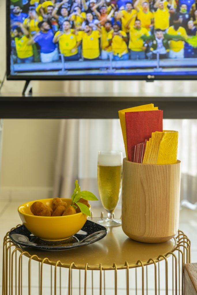 Sobre a mesinha de metal, vemos uma tigela amarela com bolinhos, uma taça com cerveja e um porta-utensílios de madeira contendo guardanapos de papel em amarelo e vermelho. 
