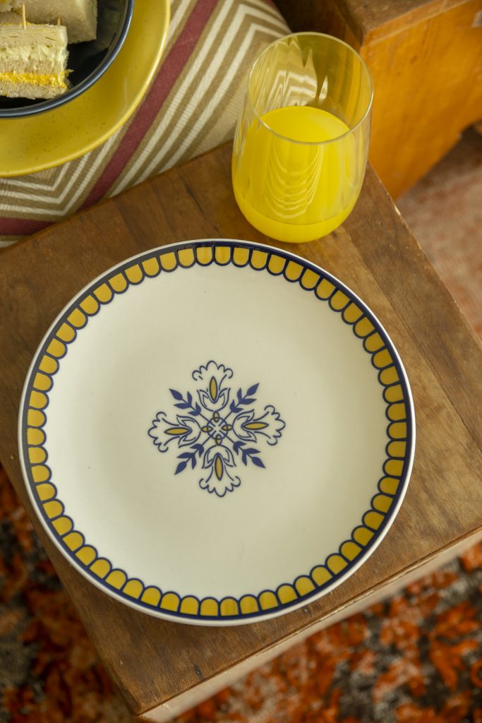 Imagem feita de cima mostra um prato branco com estampa de azulejaria em amarelo e azul. Em frente a ele, há um copo de cristal com suco amarelo. 