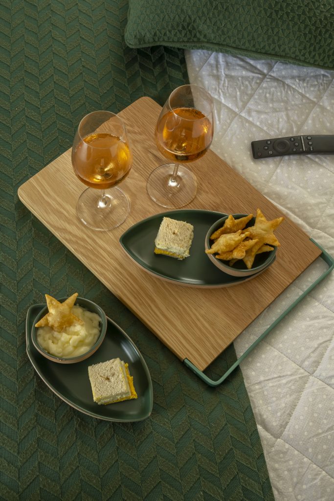 Foto feita de cima: tábua de madeira com duas taças de vinho branco, e duas tigelas de cerâmica verde sobrepostas. Do lado esquerdo, sobre a cama, vemos mais duas tigelas de cerâmica verde com petiscos.
