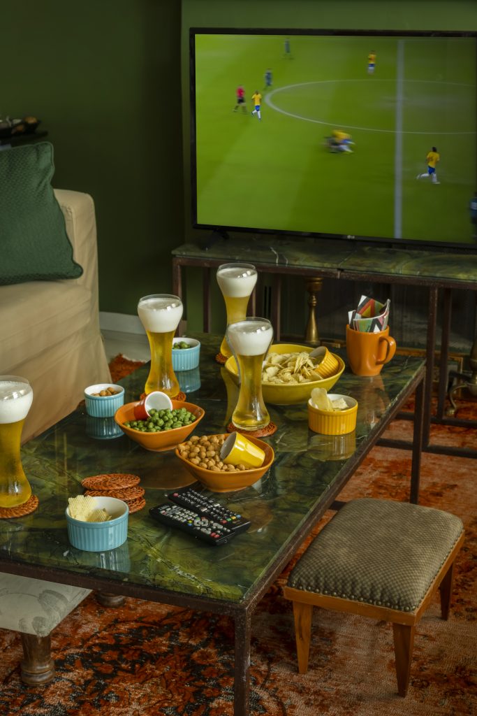 Visão parcial de uma sala de estar com mesa, sofá bege e TV de tela plana com cena de jogo de futebol com a seleção brasileira. Sobre a mesa, temos 4 copos com cerveja, e 7 tigelas, de tamanhos e cores diferentes, contendo salgadinhos.