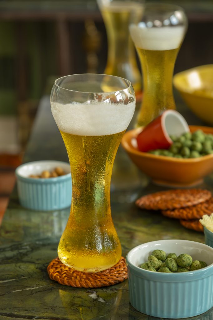Sobre a mesa, copo alto e curvo com cerveja gelada. Ao lado e no fundo, vemos tigelas de cerâmica coloridas contendo salgadinho de amendoim.
