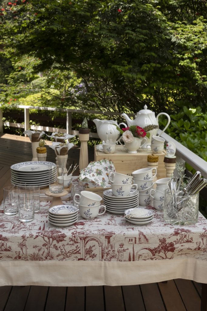 Imagem: Numa varanda com jardim ao redor, vemos uma mesa quadrada, coberta com toalha estampada em vermelho e branco. Sobre ela, temos pratos, pires e xícaras empilhados.