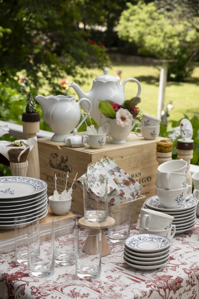 Na mesa de café da manhã de Natal, vemos uma caixa de madeira natural e, sobre ela, dois bules e um açucareiro, todos brancos. Ao redor, há pilhas de pratos, pires e xícaras de porcelana branca com estampa de ramos em azul escuro. Também há copos altos de cristal e uma toalha com estampa em vermelho. 