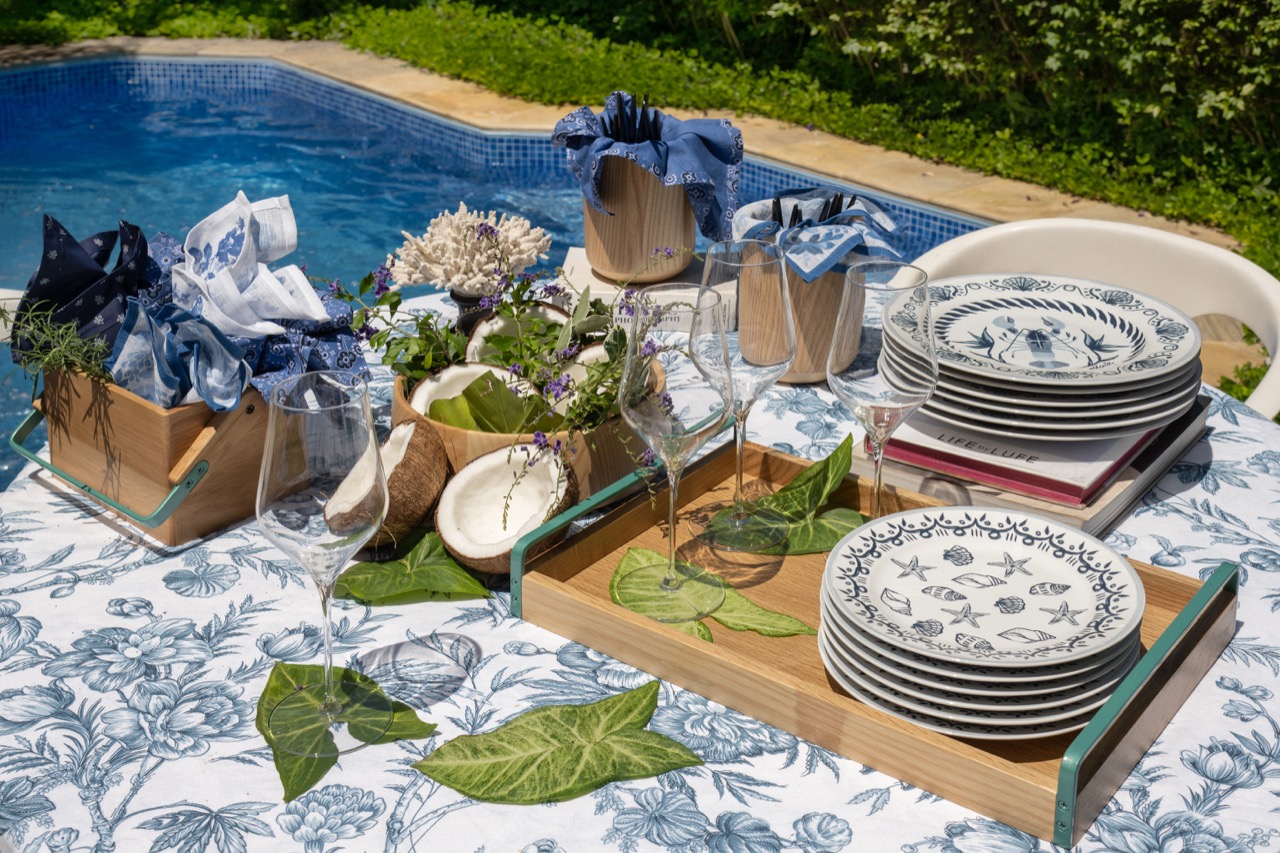 Mesa redonda, ao lado de uma piscina, com jardim ao fundo. Sobre a mesa, há pratos, taças, talheres, guardanapos e uma saladeira de madeira com metades de coco seco. 