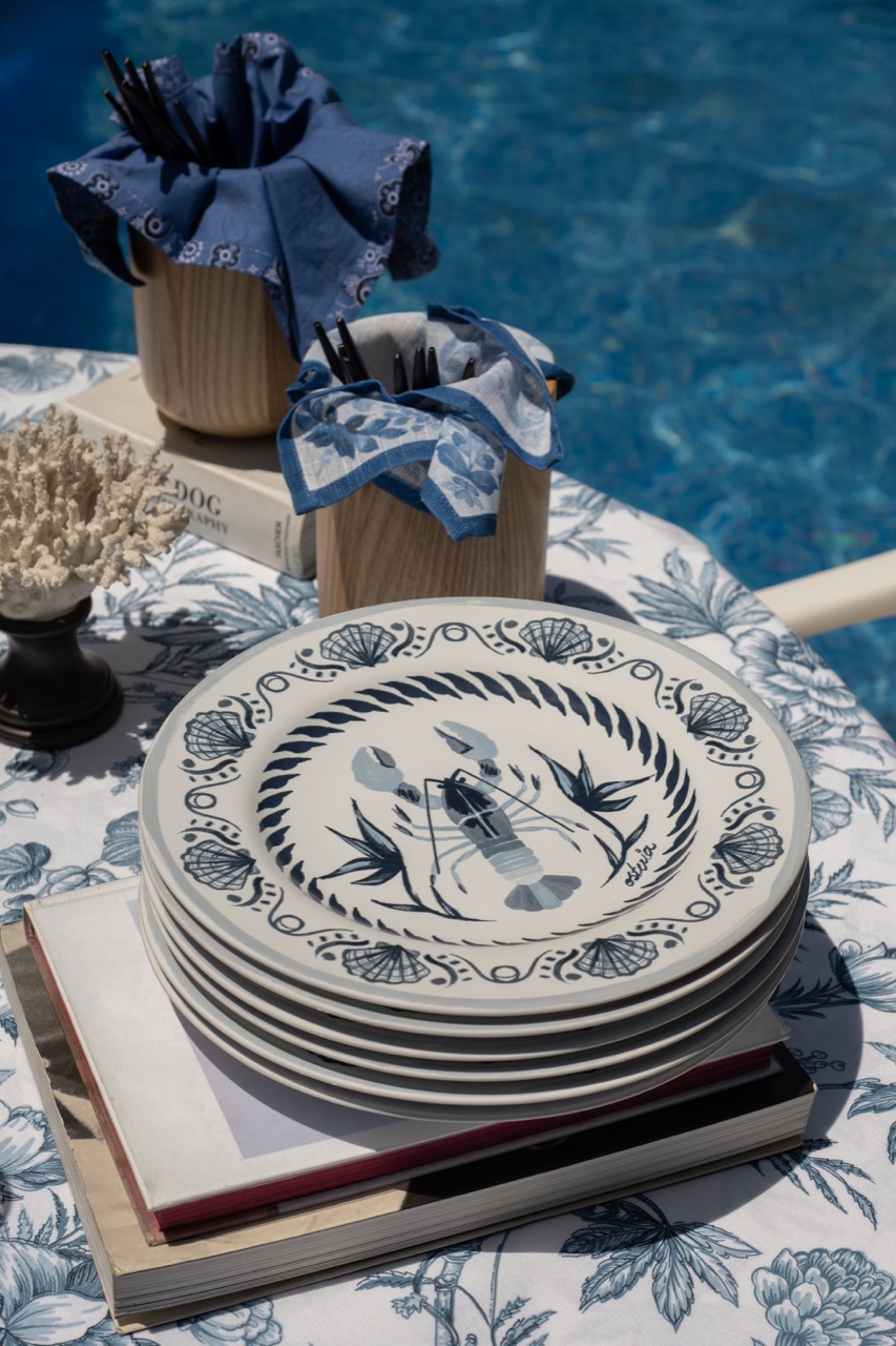 Na mesa de almoço tropical, temos uma pilha de pratos rasos, de porcelana branca, com estampa de lagosta e conhas, em azul. 
