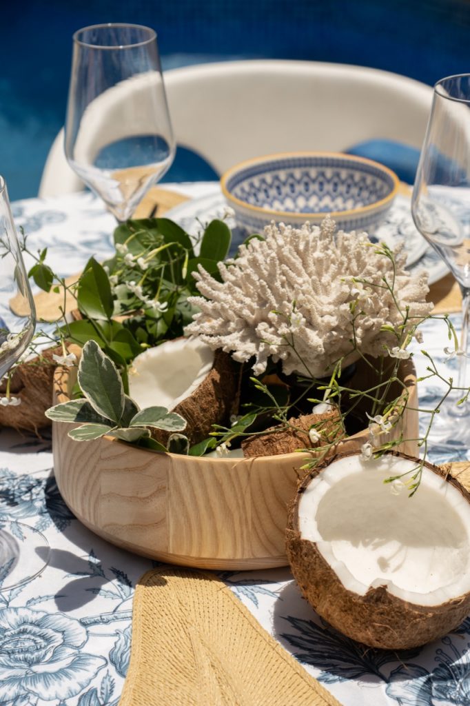 No centro da mesa posta de verão, temos uma saladeira de madeira contendo metades de coco seco, folhagens e flores miúdas. Há cocos abertos também ao lado da saladeira. 