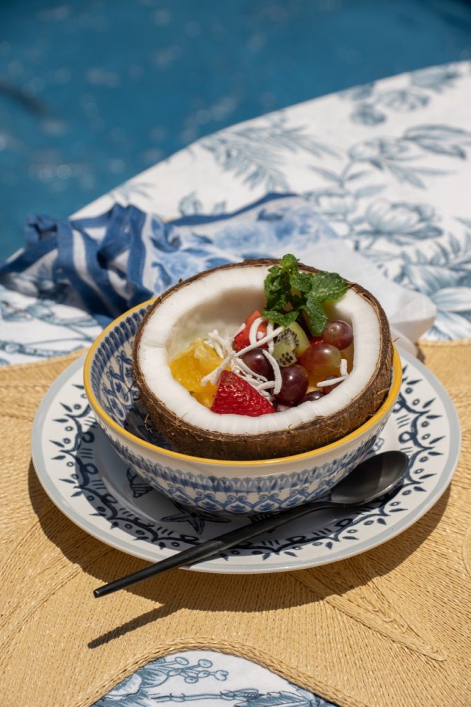 Detalhe da mesa posta de verão: dentro de uma tigela que está sobre um prato de sobremesa, vemos metade de um coco seco contendo salada de frutas. 
