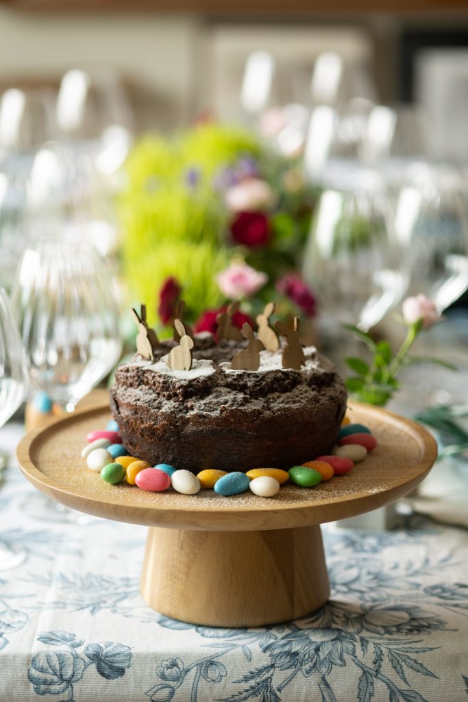 Imagem: boleira de madeira com bolo de chocolate decorado com mini ovinhos coloridos e coelhinhos para sobremesa de Páscoa.