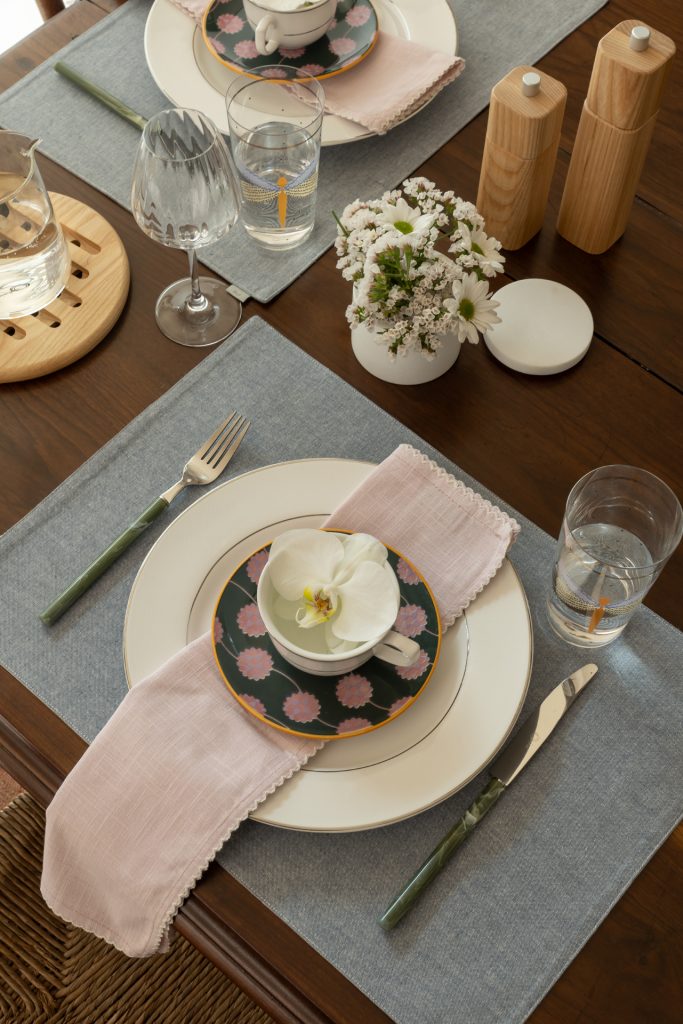 Imagem: mesa posta com pratos brancos, arranjos de flores brancas e talheres na cor verde.