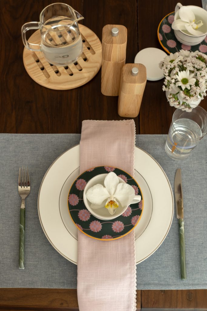 Imagem: mesa posta com pratos brancos, arranjos de flores brancas e talheres na cor verde.