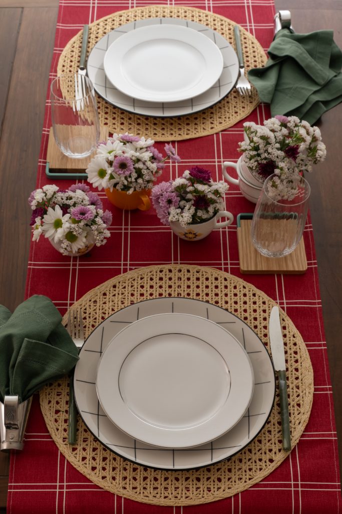 Imagem: mesa com trilho vermelho, jogo americano de palha e pratos brancos.