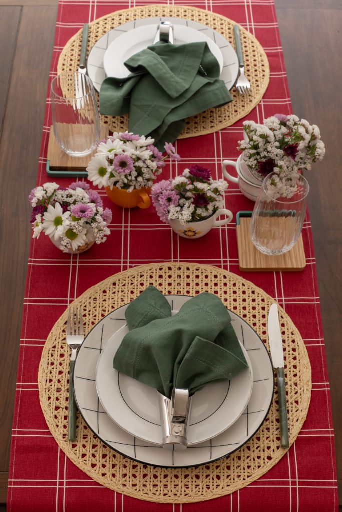 Imagem: mesa com trilho vermelho, jogo americano de palha e pratos brancos.