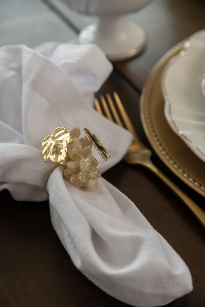 Foto aproximada de um guardanapo de tecido branco, dentro de um anel dourado enfeitado com cacho de uvas de metal e pedras rosadas, 