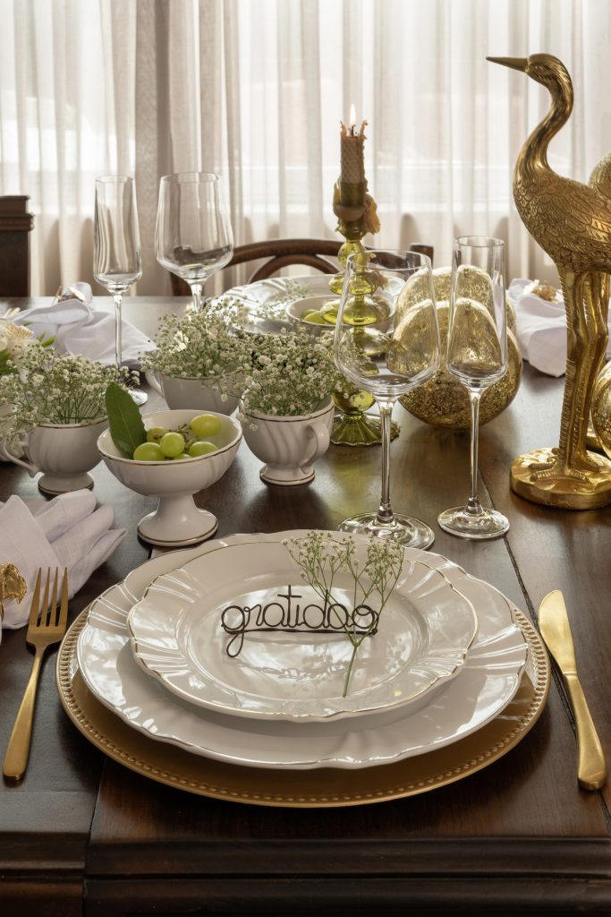 Lugar na mesa posta com prato de sobremesa sobre prato raso. Há um arame esculpido na forma da palavra "gratidão" em cima do prato menor. Os talheres são dourados.