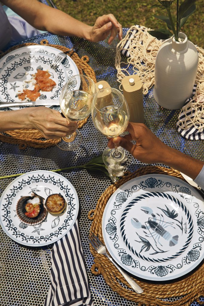 Foto feita de cima mostra parte de uma mesa posta onde há pratos brancos com estampa de lagostas e conchas em azul marinho. Vemos as mão de duas pessoas que seguram taças de cristal com vinho branco e fazem um brinde.
