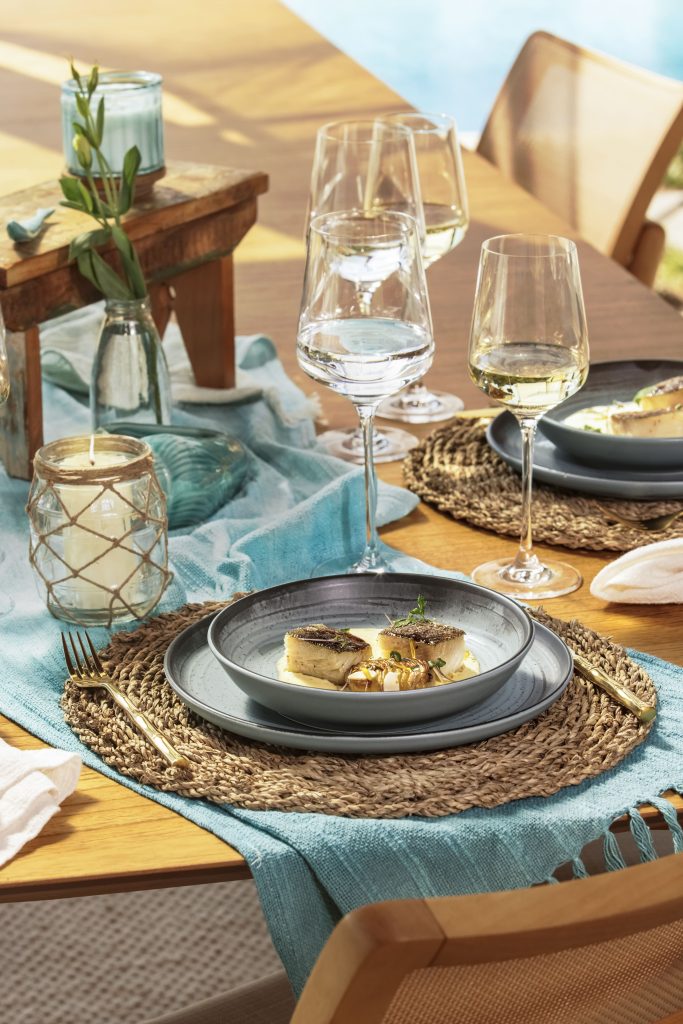 Imagem: Detalhe de mesa posta com prato fundo sobre prato raso de cerâmica, ambos em tons de cinza, sobre jogo americano de palha e caminho de mesa azul-turquesa.
