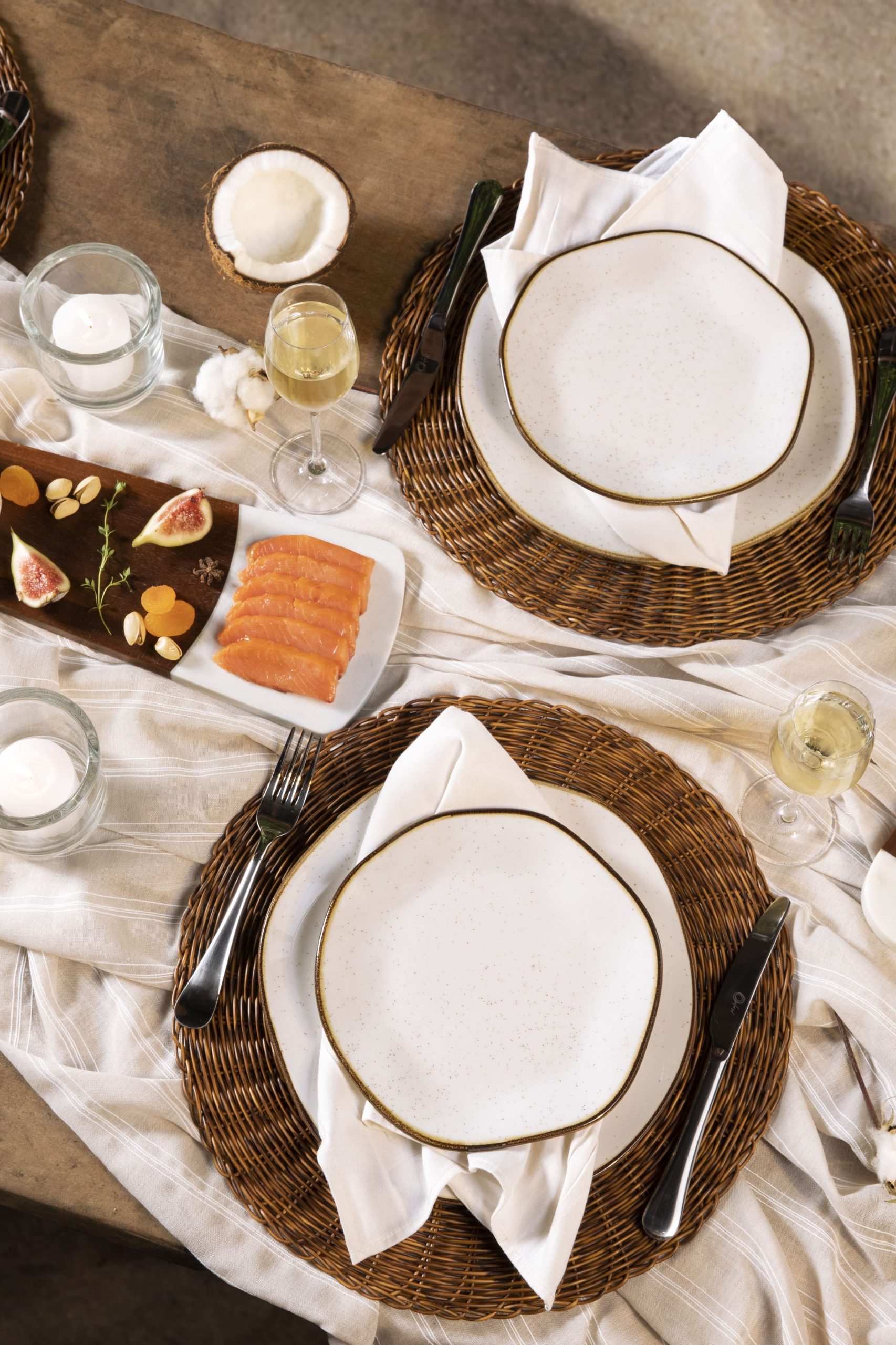 Visão parcial de uma mesa posta na praia. Sobre o tampo de madeira, vemos pratos brancos, com filete marrom nas bordas, e formas orgânicas. Há taças de cristal com espumante. No centro da mesa, vemos uma tábua com queijo e frutas, e uma vela acesa dentro de um copinho.