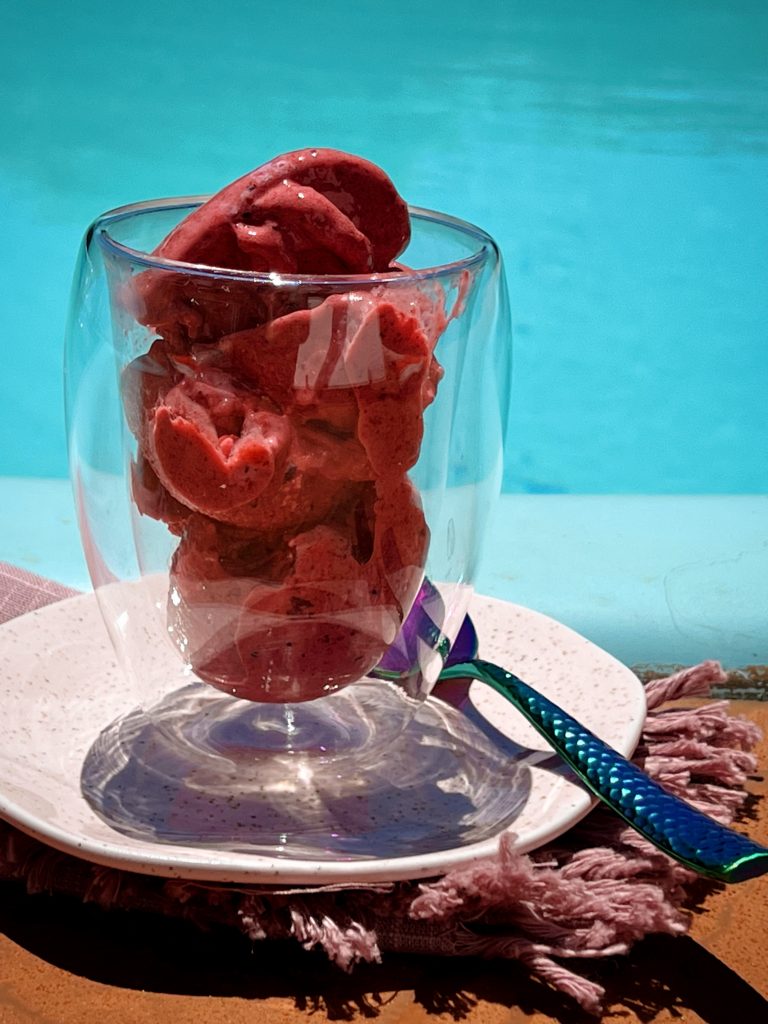 Imagem: sorvete de frutas vermelhas servido em copo transparente de parede dupla em frente a uma piscina.
