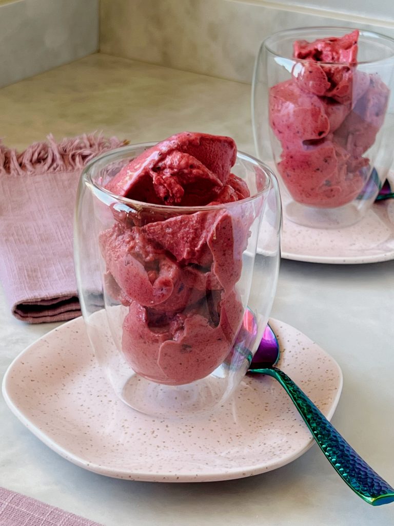 Imagem: sorvete de frutas vermelhas servido em copo transparente sobre prato rosa.