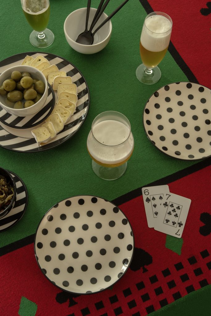Imagem: mesa com toalha para jogo com pratos de bolinhas e cartas de baralho.
