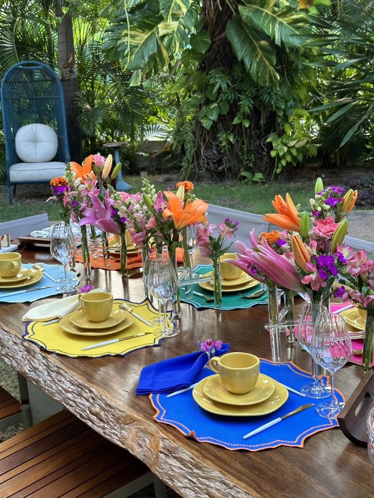 Imagem: mesa com jogo americano colorido e porcelana amarela com pratos de sobremesa, xícara e pires. No centro da mesa arranjos altos de lírios laranja e flores do campo rosa.