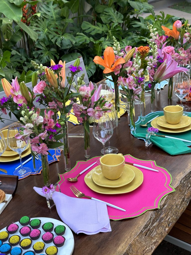 Imagem: mesa com jogo americano colorido e porcelana amarela com pratos de sobremesa, xícara e pires. No centro da mesa arranjos altos de lírios laranja e flores do campo rosa.