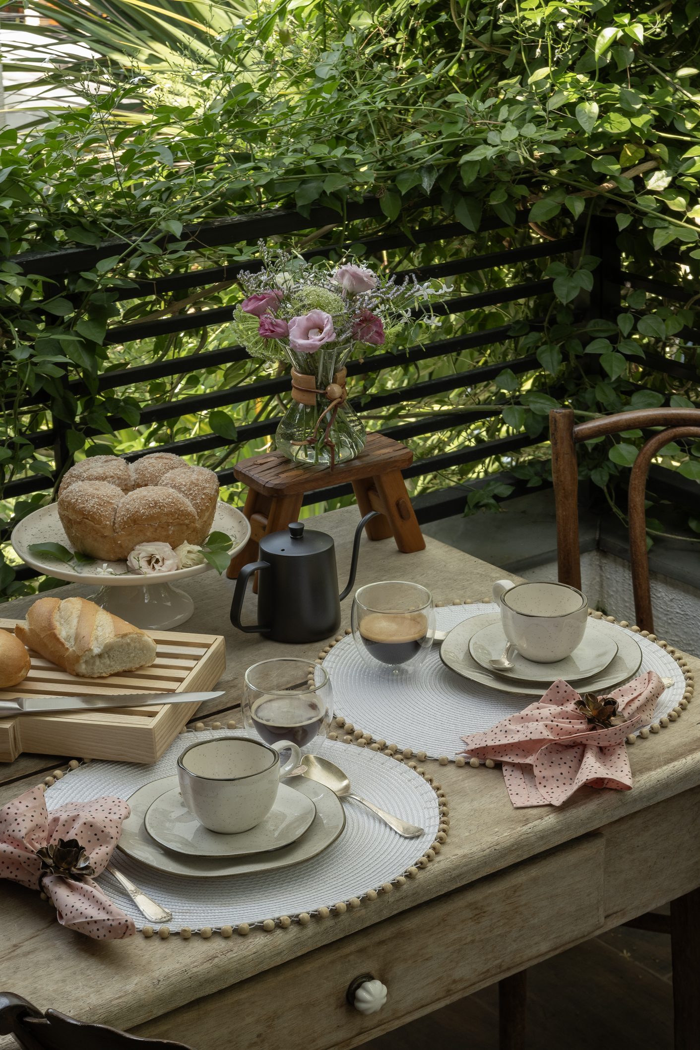 Imagem: mesa posta em varanda com pratos de sobremesa, xícaras e pires, tábua de pão e copos baixos de cafezinho. 