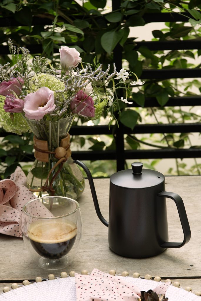 Imagem: copo de vidro com café e jarra bico de ganso sobre mesa.