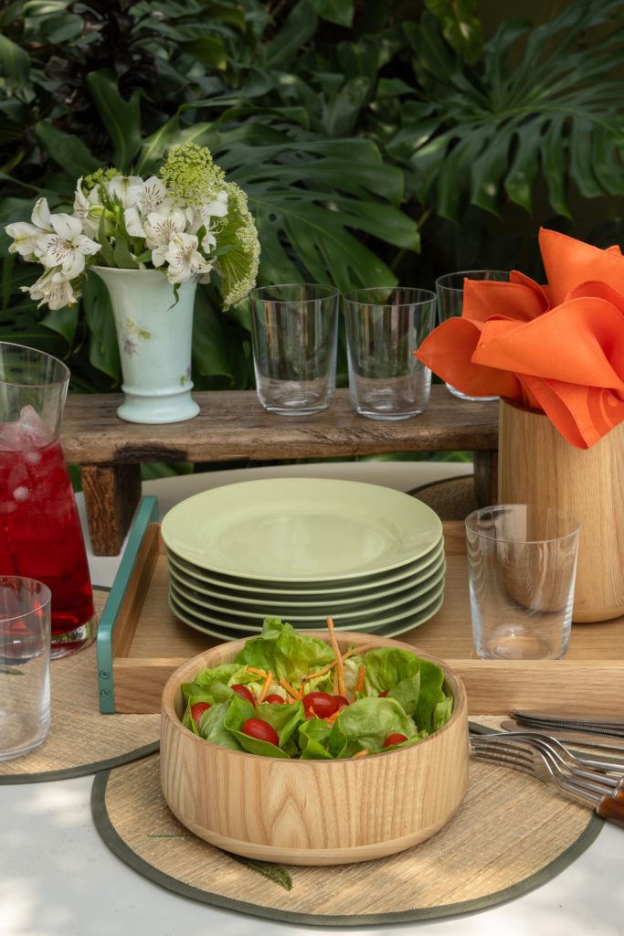 Imagem: buffet com pratos verdes da Oxford e cumbuca de madeira com salada de folhas. Copos de cristal servidos com suco vermelho. 