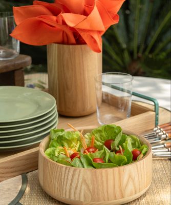 Imagem: tigela de madeira da Oxford servida com salada de flores e tomate. Porta-utensílios de madeira com guardanapos de pano na cor laranja.