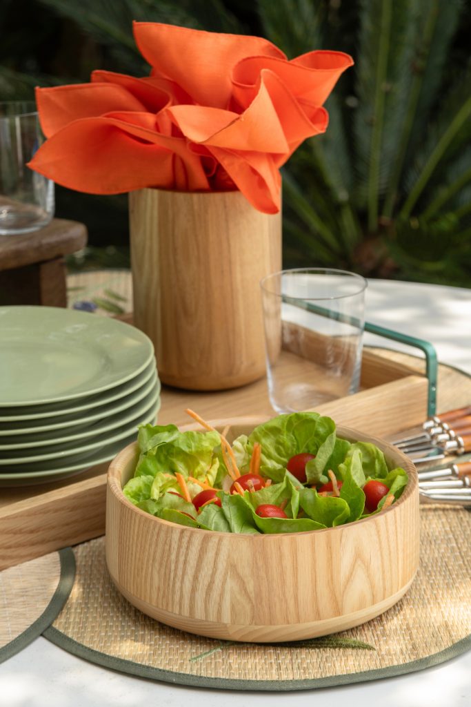 Imagem: tigela de madeira da Oxford servida com salada de flores e tomate. Porta-utensílios de madeira com guardanapos de pano na cor laranja. 