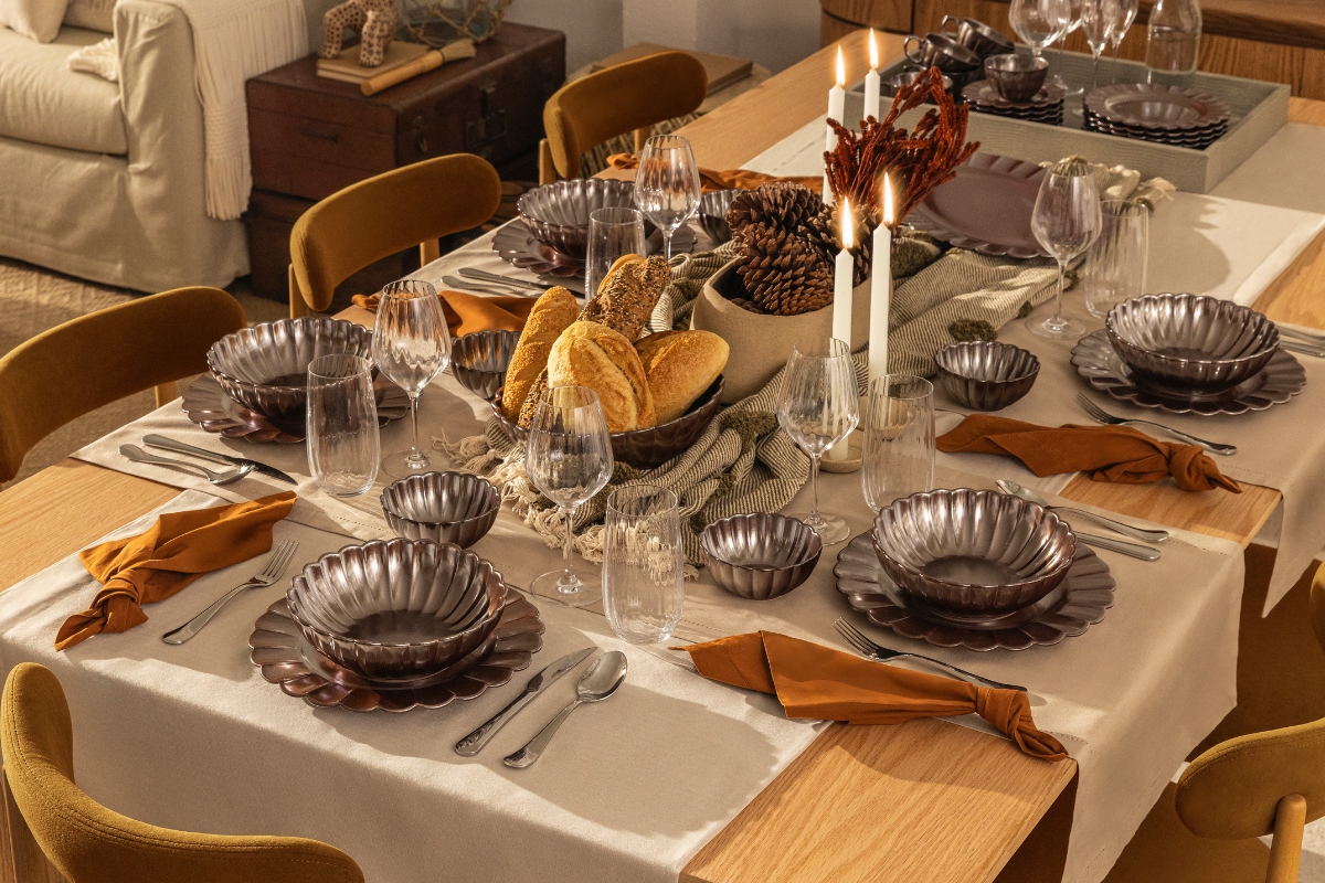 Imagem: Mesa posta com louças Oxford em tom de marrom com bordas onduladas e acabamento brilhoso, acompanhadas de taças, guardanapos alaranjados e decoração com pães, pinhas e velas brancas acesas.